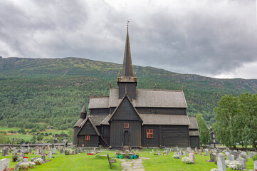 Fototapeta na wymiar Lom viking medieval stave church in Lom Norway. Old wooden walls and tower, green lawn and grey graveyard tombstones, mountain slope and cloudy sky on the background