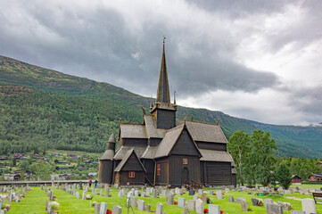 Fototapeta na wymiar Lom viking medieval stave church in Lom Norway. Old wooden walls and tower, green lawn and grey graveyard tombstones, mountain slope and cloudy sky on the background