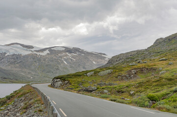 road in the Norwegian mountains, summer landscape with snow. Green and grey rocky slopes, mossy stones, cloudy sky, grey water of the mountain lake
