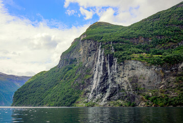 View of Geirangerfjord and the Seven sisters waterfall in Norway from the water.  Blue sky with clouds, green mounain slopes with sunshine and shadows on the forest