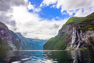 View of Geirangerfjord and the Seven sisters waterfall in Norway from the water.  Blue sky with clouds, green mounain slopes with sunshine and shadows on the forest