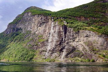 View of Geirangerfjord and the Seven sisters waterfall in Norway from the water.  Blue sky with clouds, green mounain slopes with sunshine and shadows on the forest