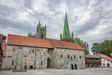 Fototapeta na wymiar Nidarosdomen gothic cathedral and bishop`s palace in Trondheim, Norway. Blue and lilac sky with grey clouds, grey medieval stone walls and bell towers, green trees, cloudy sky