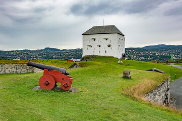 Kristiansten fortress in Trondheim, Norway. White walls with gun ports, green grass on the ramparts, old cannon on the stone wall, grey cloudy sky - Powered by Adobe