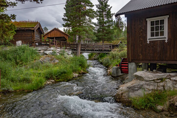 Fototapeta na wymiar Old wooden watermill in Norway. White water of the mountain stream, tiny wooden buildings, grey stones on the green grass, cloudy sky