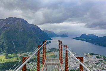  Rampestreken lookout point on the trail to Nesakla summit in Norway.  Cloudy sky, grey water of Romsdalsfjord, dark mountains, town of Andalsnes down there