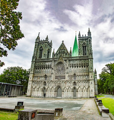 Fototapeta na wymiar Nidarosdomen gothic cathedral in Trondheim, Norway. Blue and lilac sky with grey clouds, grey medieval stone walls and bell towers, figures of the saints, green grass and trees