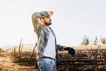 Young worker farmer man tired after work, next to his farmland and looking at his plantations. 