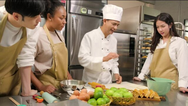 Cuisine course, senior male chef in cook uniform teaches young cooking class students to knead pastry dough and prepare ingredients for bakery foods, fruit pies in restaurant stainless steel kitchen.