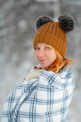 Close up portrait of a young Caucasian red hairy woman in the winter snowy forest. Young woman wrapped in a warm white-blue plaid. Winter leisure in the park.