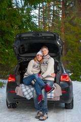A married couple enjoying beautiful winter weather in the open trunk of a car in the winter snowy forest. Happy caucasian family enjoying the romantic outdoor date, posing and looking into the camera.