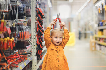 Obraz premium Cute girl child in mustard dress in hardware store, child plays with pliers, chooses tools in large hypermarket, child safety