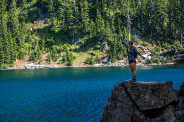 Adventurous athletic woman standing on a bolder at an alpine lake with her arms open wide, smiling at the camera.