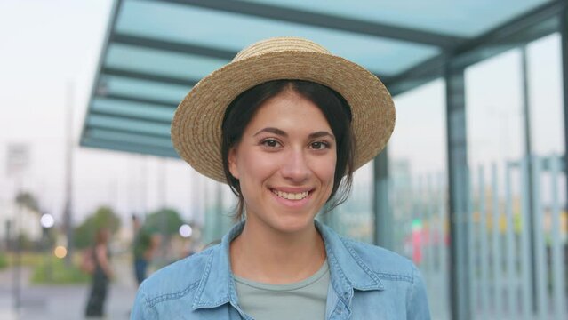 Close up of young girl with brown hair in hat. Beautiful woman turns her head, touch hair and smiles at camera. Female in sunny day on bus station outdoor. Pretty joyful good-looking sunshine portrait