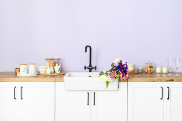 Sink with beautiful flowers and kitchenware on counters near lilac wall