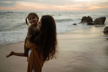 Lovely baby daughter embrace mother on the beach at sunset. Summer family vacation by the sea