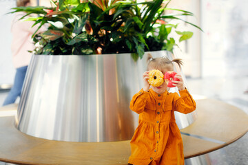 Toddler girl eats donut in supermarket on bench, snack with pastries shopping. Cute child girl in long sleeve mustard dress plays with yellow and pink donuts
