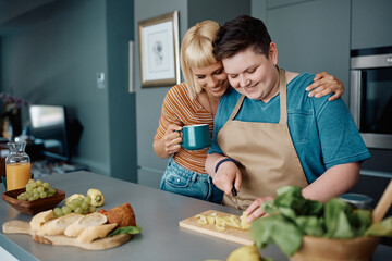 Happy lesbian drinks coffee and embraces her girlfriend who is preparing food in kitchen. - obrazy, fototapety, plakaty
