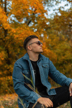 Fashion young man model hipster in fashionable denim clothes with jeans jacket and sunglasses sits in autumn colored park with golden leaves. Guy rest outdoors