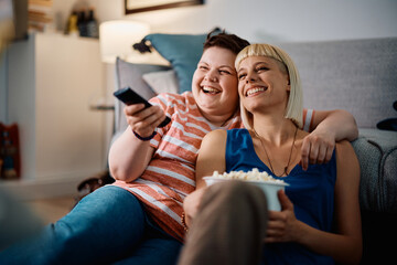 Cheerful gay women have fun while watching TV and eating popcorn at home.