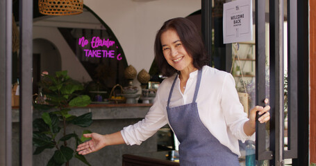 Asia latin adult female SME worker stand pride at front door work smile laugh look camera in new store small coffee shop. Happy casual day enter pub bar cafe warm hand sign for food drink industry.