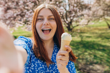 Happy cheerful young redhead woman in fashion dress posing outside with ice cream. Woman taking selfie and holding waffle cone of sweet food on nature background, summer time