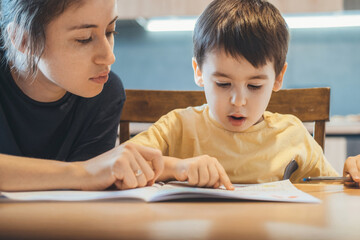 Caucasian little boy and his mother sitting together at table and reading book, mother teaching her...