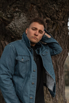 Fashionable handsome young man with hair in a fashion blue jeans jacket and black T-shirt stands and poses near a tree in the countryside outdoors