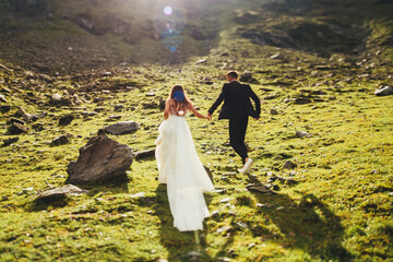 Back view of a groom in a suit and a bride in a white dress are running on the mountain hills,...