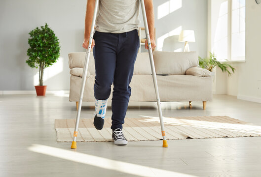 Closeup crop of unhealthy injured man with bandage on foot walk on crutches at home. Unwell guy have splint or sling on leg for recovery. Injury and trauma rehabilitation. Healthcare concept.