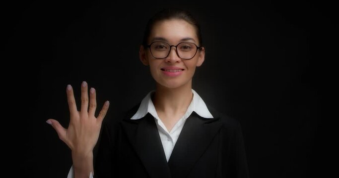 Positive asian woman in glasses and business clothes, smiling, shows five fingers with her right hand. Businesswoman counts with her fingers on her hand. Isolated on a black background.