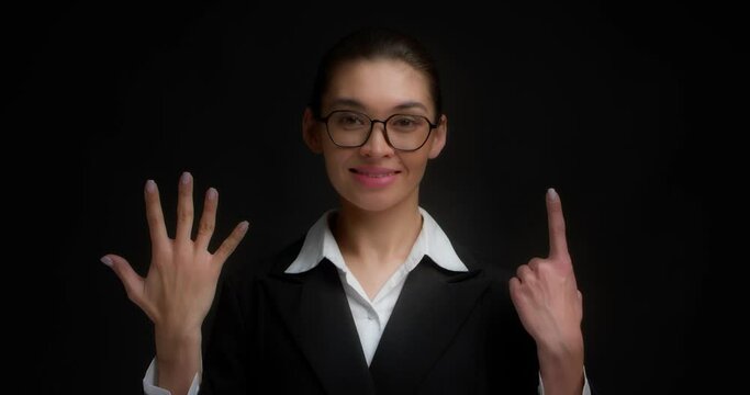 Positive asian woman in glasses and business clothes, smiling, shows six fingers with her hand. Businesswoman counts with her fingers on her hand. Isolated on a black background.