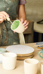 Fototapeta na wymiar Close-up of girl painting clay mug with glaze. Woman coloring pottery in workshop with a paintbrush. Painter in green apron glazing clay pot