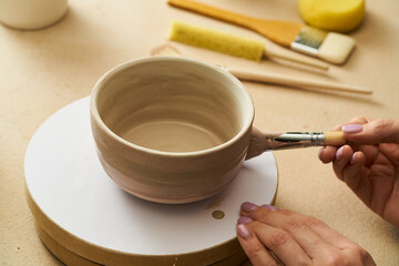Close-up of girl painting clay mug with glaze. Woman coloring pottery in workshop with a paintbrush. Painter in green apron glazing clay pot. - 502901687