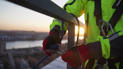 Man attaches rope with carabiner on roof steel fence. Industrial climber fastens safety equipment for climbing down building at sunset closeup