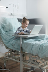 Happy ill little child enjoying video games on laptop while resting in hospital bed. Smiling sick little girl playing games on modern computer laptop while in pediatric clinic recovery ward room.