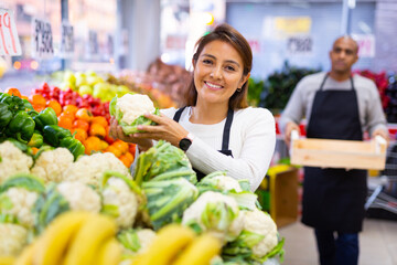 Salesgirl standing near fresh cauliflower and vegetables in grocery store