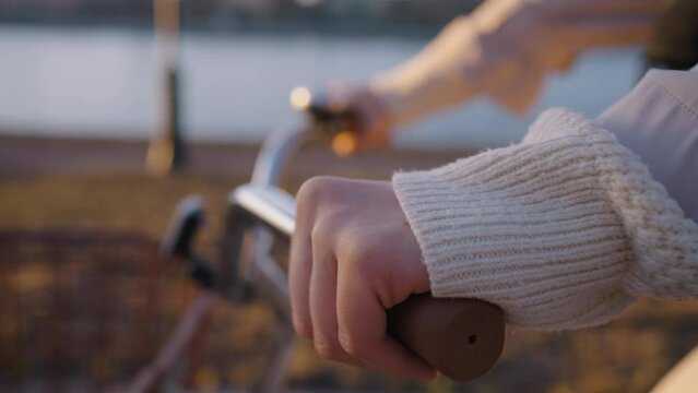 Hands of woman riding vintage bicycle with basket along alley with bare trees past river. Bike rides along embankment at golden hour closeup