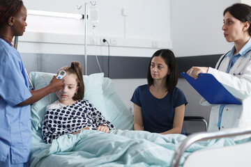 Pediatric clinic nurse measuring ill body temperature using modern thermometer while in healthcare pediatric hospital patient recovery ward room. Pediatrist writing down analysis results.