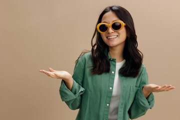 Enjoying Korean young woman in khaki green shirt stylish eyewear raise palms up hold invisible objects posing isolated on over beige pastel studio background. Cool fashion offer. Sunglasses ad concept