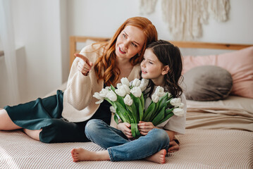 Happy beautiful affectionate mother, with a bouquet of spring flowers, hugs her daughter at home on the bed, enjoys the moment of surprise, celebrates Mother's Day.