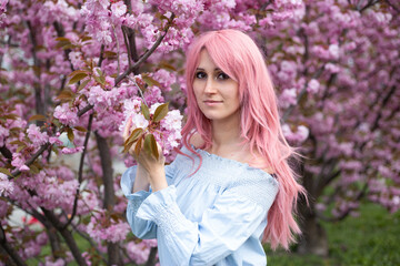 Fototapeta premium beautiful woman in pink wig near sakura blooming tree. Pink flowers, spring and youth concept. Stylish girl smiling. Blooming garden with cherry-tree. Copyplace, place for text.