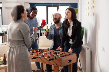 Diverse group of colleagues having fun with foosball table game, enjoying after work drinks and...