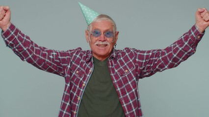 Cheerful positive surprised senior man in cone festive hat cap, dancing, having fun, celebrating birthday party event holiday, anniversary. Mature old grandfather alone on gray studio wall background