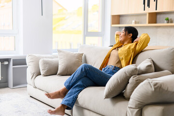 Fototapeta premium Charming young African-American woman resting, peaceful and serene multiracial lady looking at the camera sitting on the sofa in cozy living room, spends weekend at home