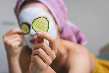 Young adult woman in cosmetic facial mask holds slices of cucumber in front of face.