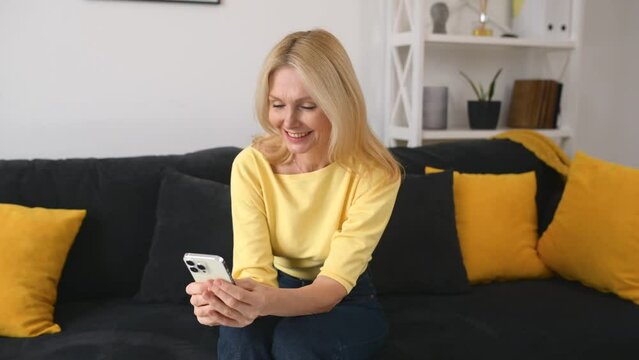 Serene 50s middle-age woman using smartphone sitting on the sofa at home, smiling carefree female web surfing, scrolling news feed, looking through photos
