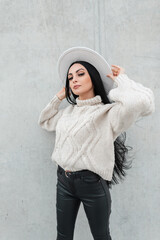 Fashion pretty woman with glasses in casual urban clothes with white knit sweater, leather pants and white hat poses near a gray wall on the street