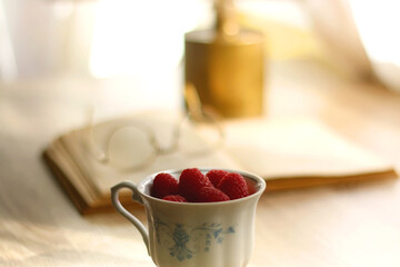 Porcelain cup filled with raspberries, open book, reading glasses and vase with flowers on the table. Selective focus.