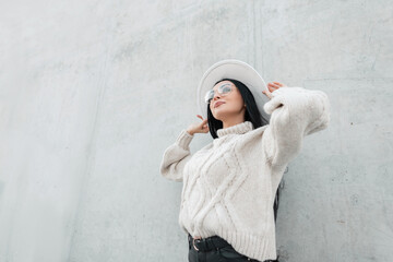 Beautiful fashionable young woman with glasses in a stylish vintage comfortable sweater is wearing a hat and standing near a gray concrete wall on the street